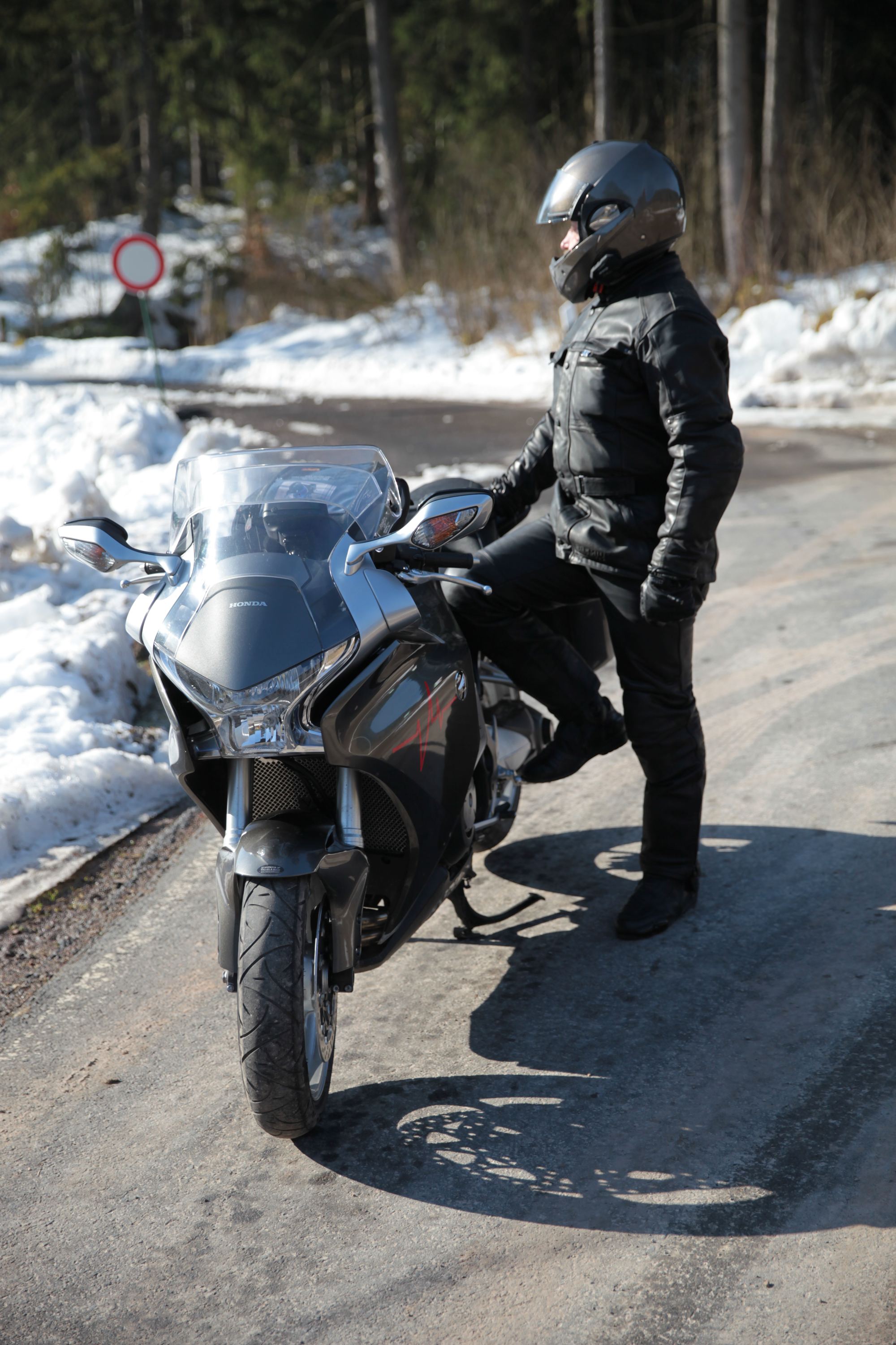 Fahrer in schwarzer Lederkleidung neben einer grauen Honda auf geräumter Straße, links Schneewälle und ein rot umrandetes Rundschild Fahrer in schwarzer Lederkleidung neben einer grauen Honda auf geräumter Straße, links Schneewälle und ein rot umrandetes Rundschild