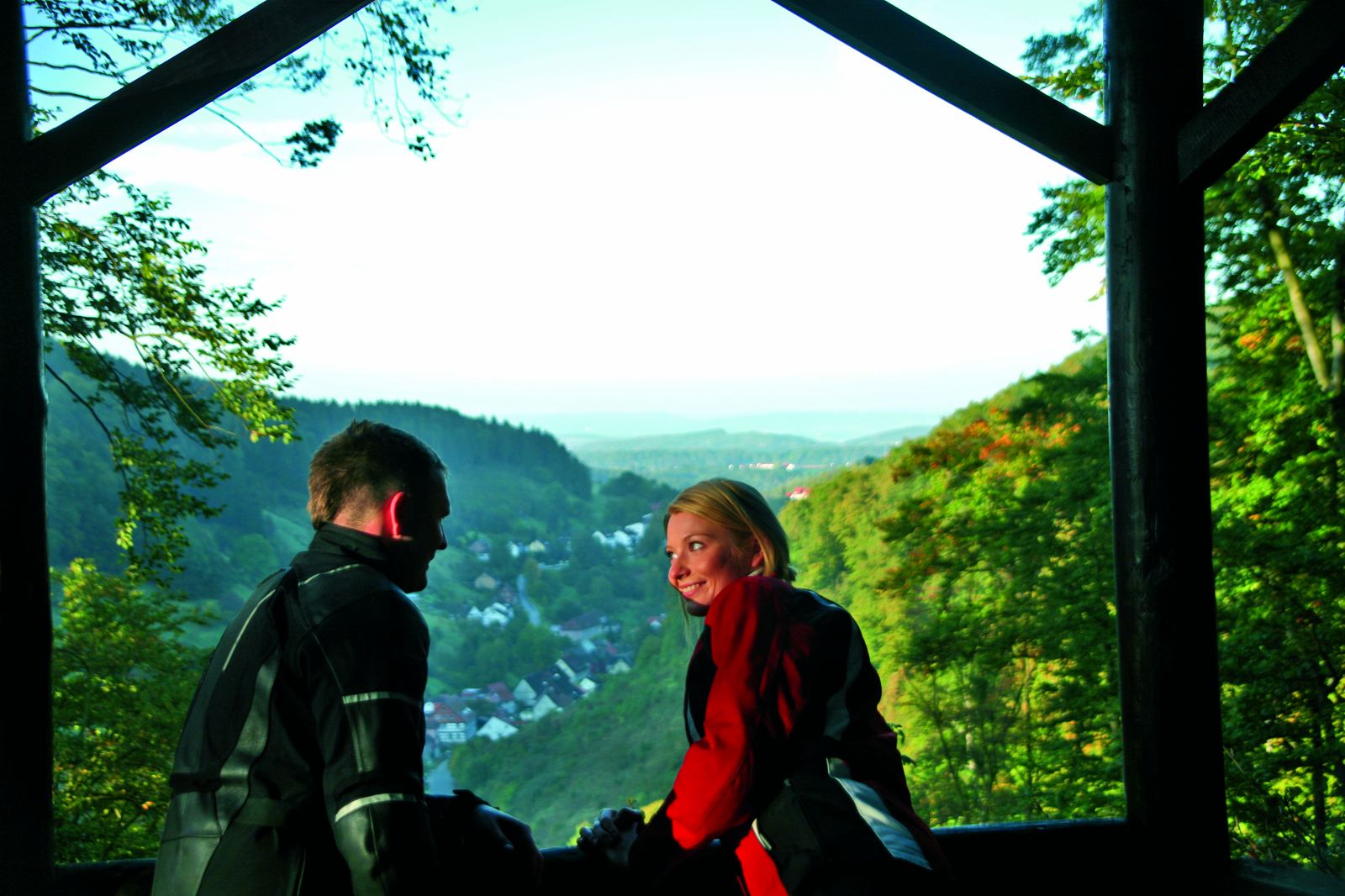 Paar in Motorradkleidung in einer Holzhütte am Hang, darunter ein langgestrecktes Dorf im Tal Paar in Motorradkleidung in einer Holzhütte am Hang, darunter ein langgestrecktes Dorf im Tal