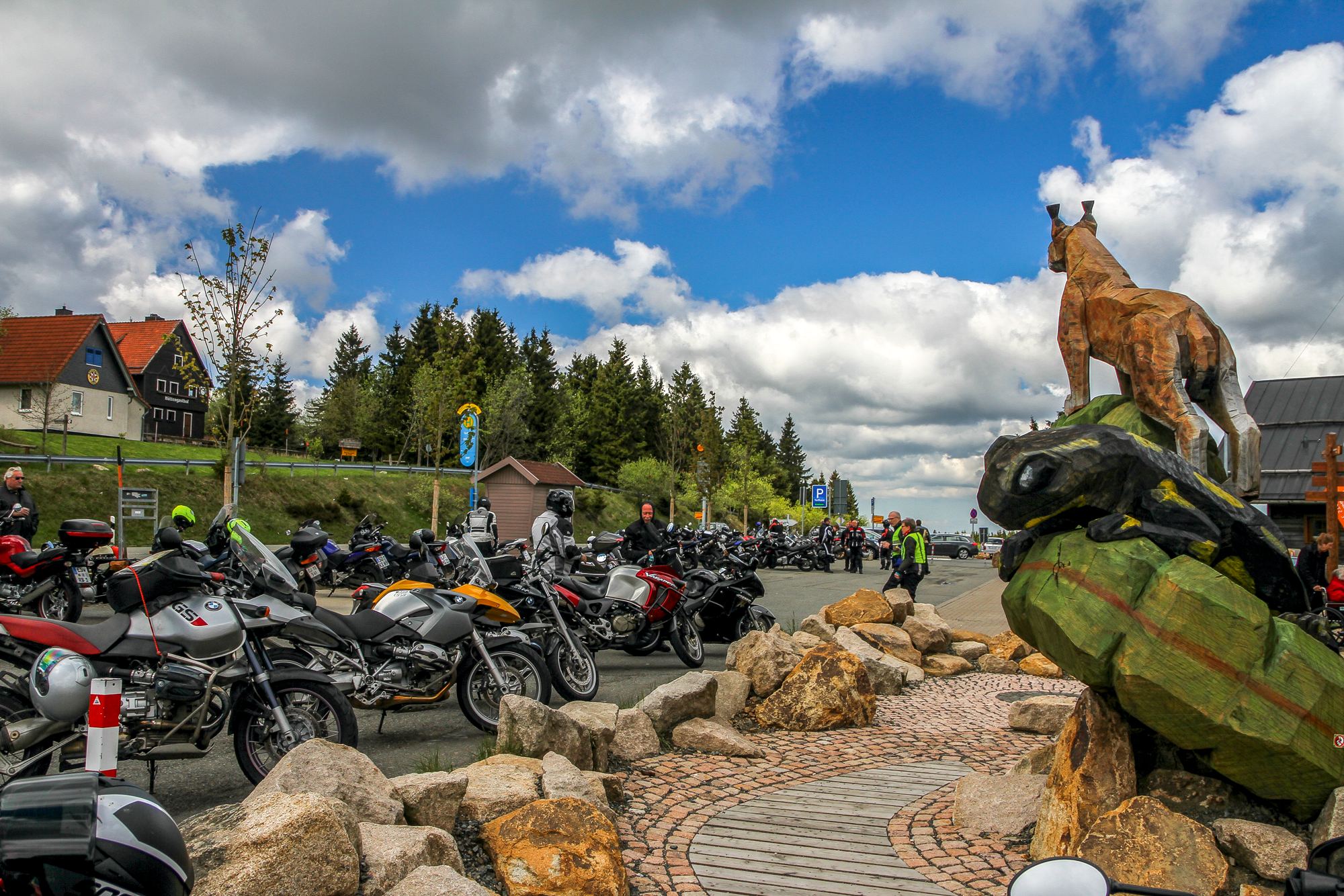 Kettensägen-Skulptur eines Luchses auf einem bemalten Holzfelsen, dahinter ein Parkplatz voller Motorräder Kettensägen-Skulptur eines Luchses auf einem bemalten Holzfelsen, dahinter ein Parkplatz voller Motorräder